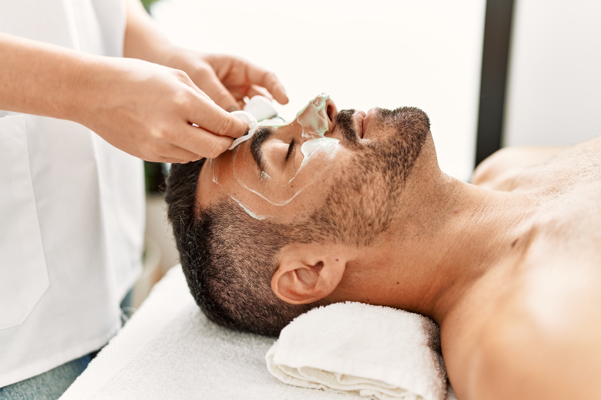 Young hispanic man having facial mask treatment at beauty center